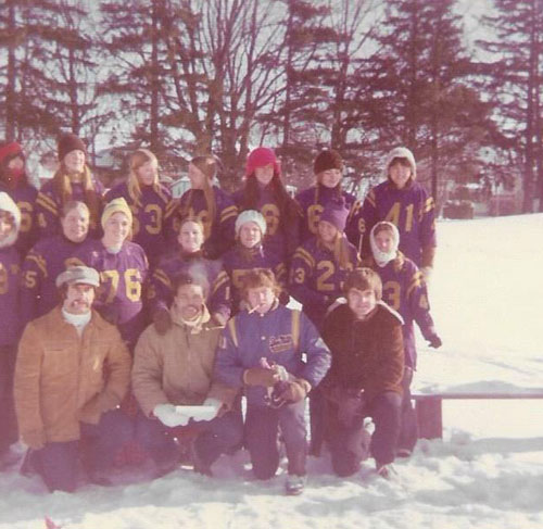 Young female football players posed in a group shot in the snow, wearing purple and gold jerseys. A few men posed in the front row, wearing street clothes.